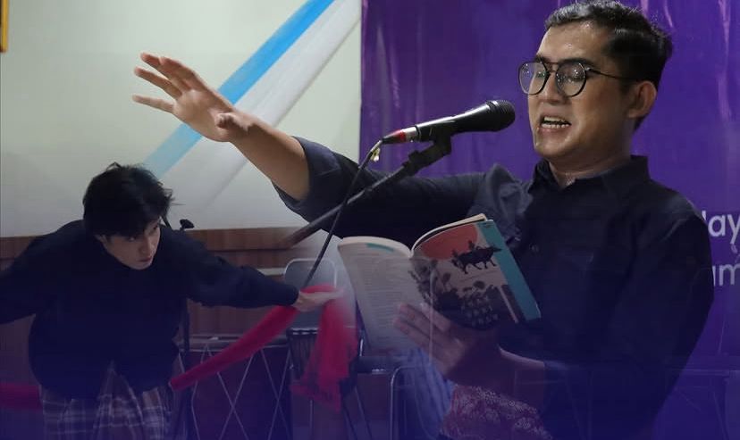 Seorang pembaca sastra berkacamata membacakan karya di depan mikrofon